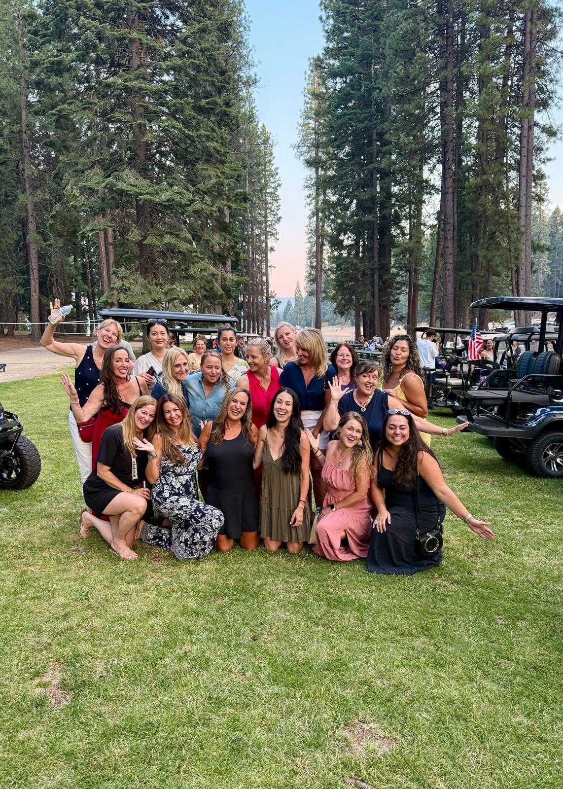 Large group of women posing and smiling on a lawn surrounded by tall pine trees