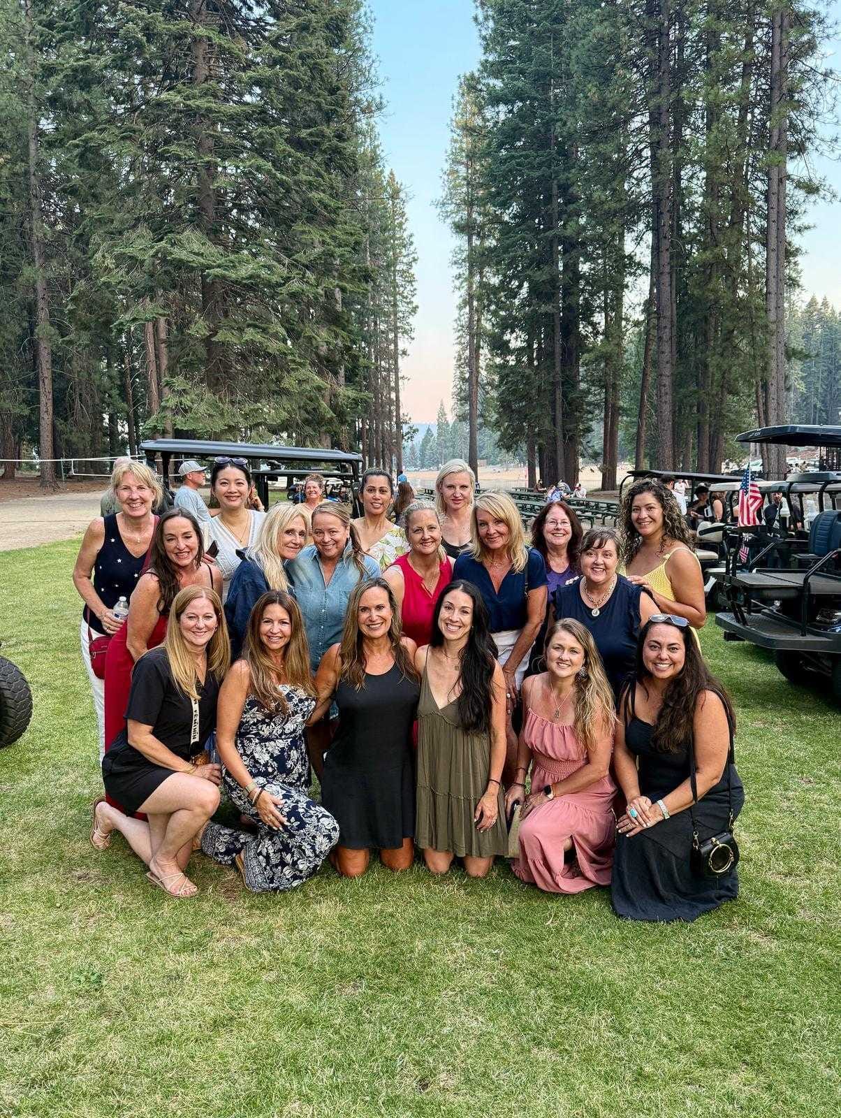Large group of women posing together on a grass lawn surrounded by tall pine trees