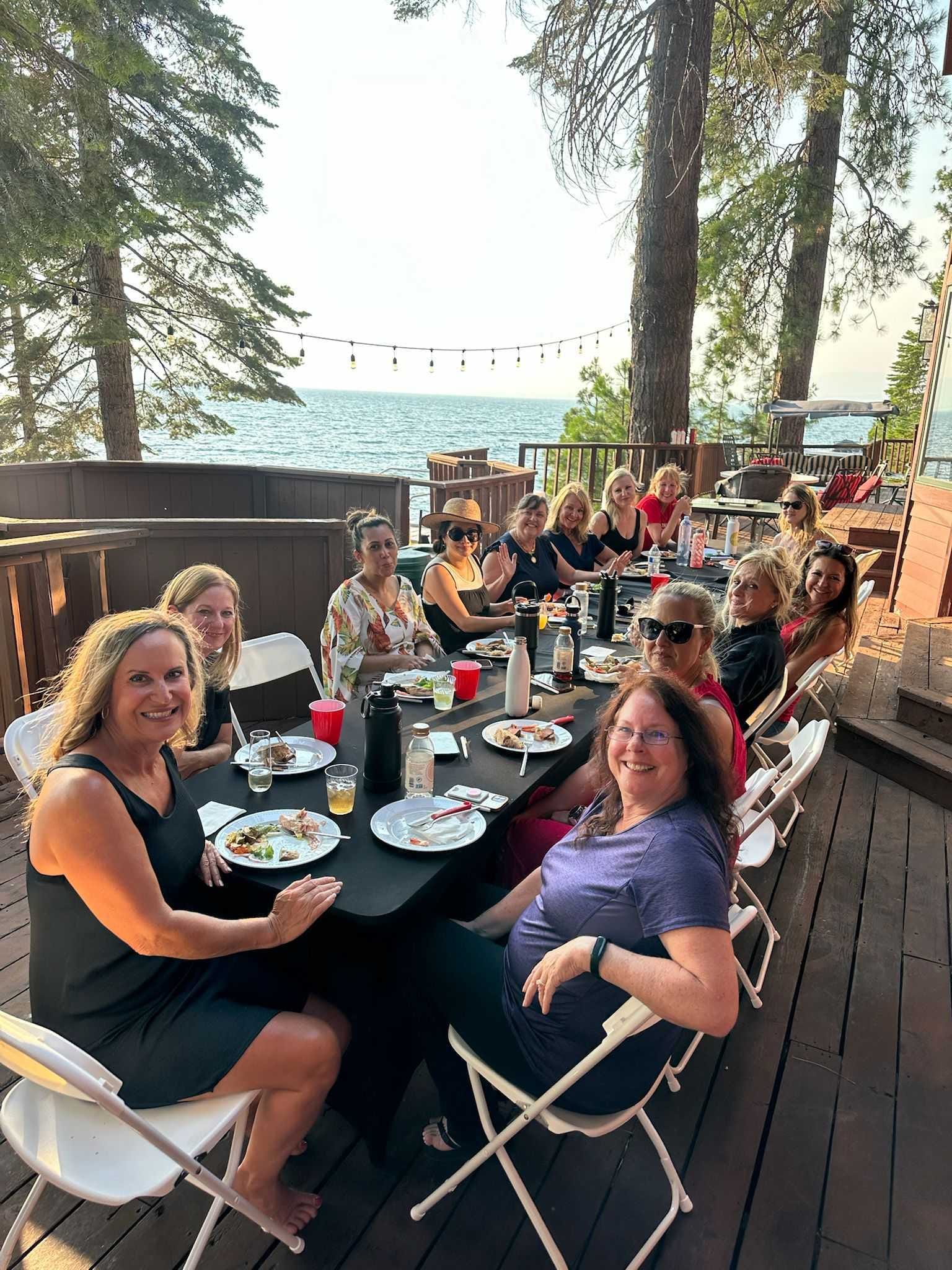Group of women dining together at an outdoor lakeside deck surrounded by pine trees