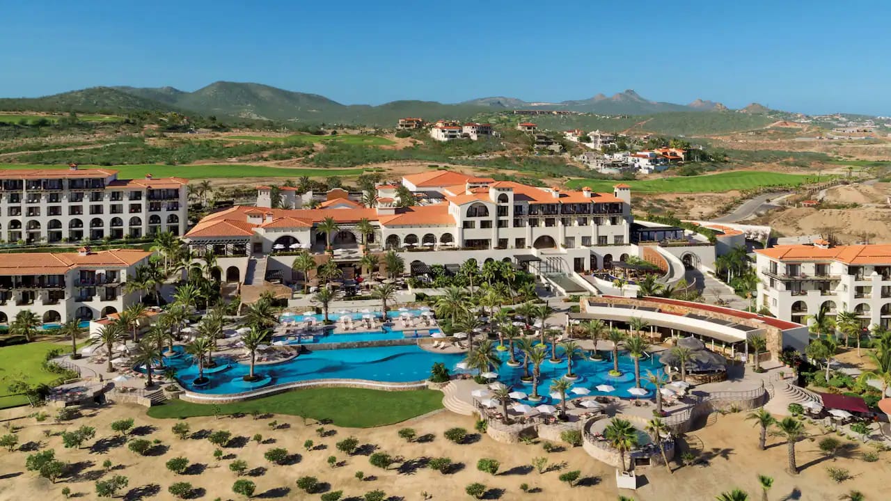 Aerial view of a luxury resort with pools, palm trees, and terracotta-roofed buildings against green hills